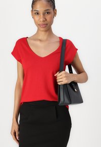 Červený top s krátkým rukávem a výstřihem do V, doplněný černou přiléhavou sukní. V ruce je držen černý handbag s elegantním designem a zlatým akcentem.