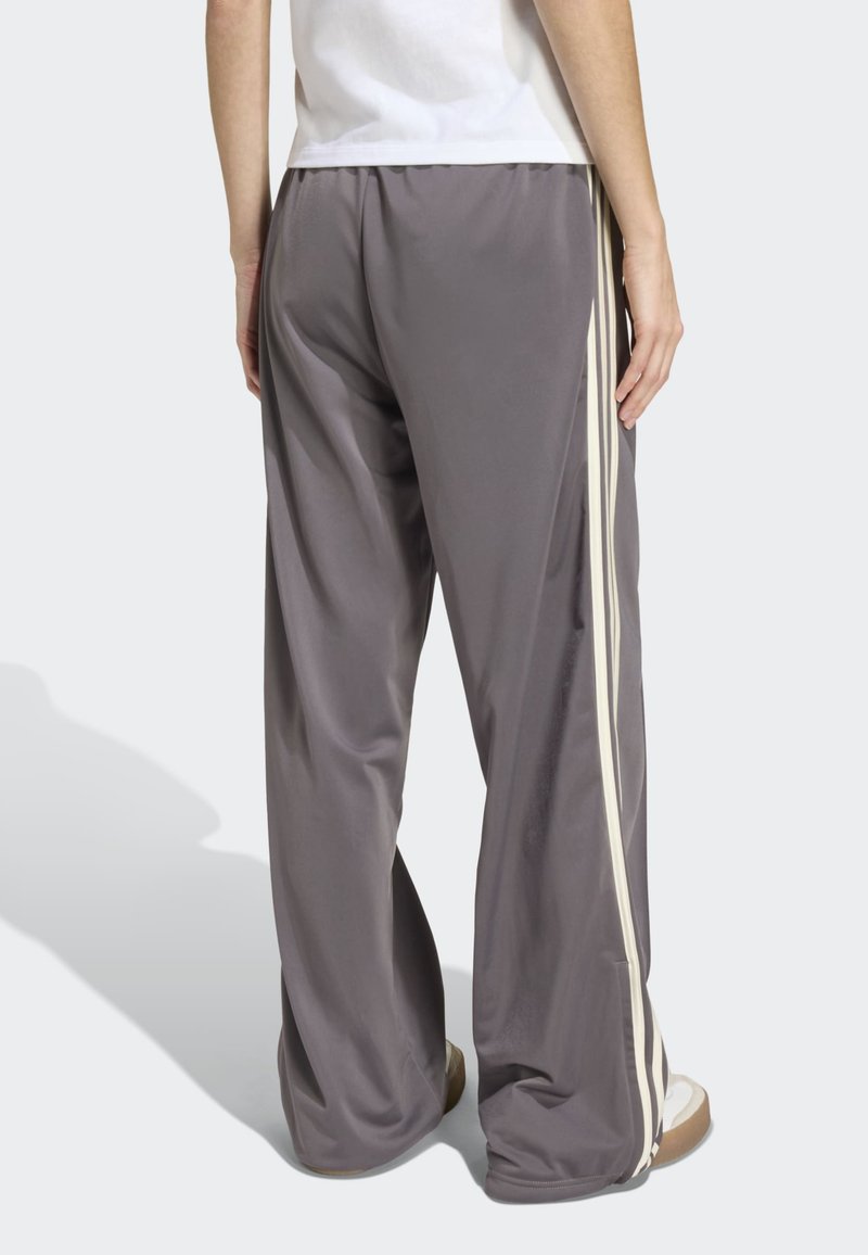 Personne portant un pantalon de survêtement gris ample avec des bandes latérales beiges et des baskets blanches, debout, dos à l'appareil photo.