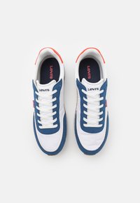 Sneakers Levi's z białą skórzaną cholewką, niebieskimi zamszowymi akcentami, czerwonymi paskami na pięcie, białymi sznurówkami oraz teksturowaną gumową podeszwą.