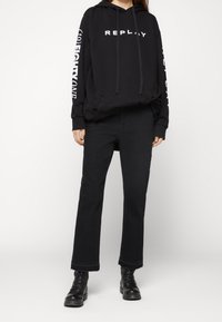 Sudadera con capucha negra con el texto "REPLAY" en el pecho y "EIGHTYONE" en la manga, combinada con unos jeans negros de pierna recta y botas negras.