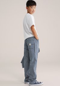 Poiss valges t-särgis, triibuliste siniste pükstega ja mereväe tennistega seisab käed taskutes, vaadates üle õla lihtsal taustal.