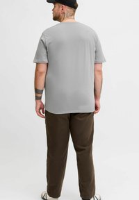 Man with tattoos wearing gray t-shirt, brown pants, black cap, and white sneakers stands facing away on plain background.