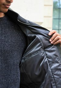 Schwarze Zip-Jacke mit glattem Innenfutter, ausgestattet mit einer Innentasche und einem gerippten Kragen. Getragen über einem strukturierten dunklen Pullover.