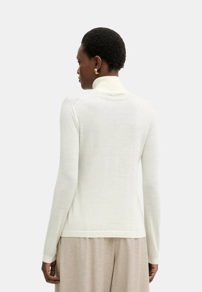 Donna con capelli corti che indossa orecchini a cerchio dorati, un maglione dolcevita bianco a maniche lunghe e pantaloni beige, vista da dietro.