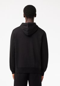 Homme aux cheveux courts et bouclés portant un sweat à capuche noir uni et un pantalon noir, debout de dos sur un fond blanc.