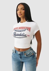 Biały skrócony t-shirt z wielokolorowym graficznym napisem. Jasnoniebieskie obcisłe dżinsy z przetarciami, z miedzianymi guzikami i przeszyciami.