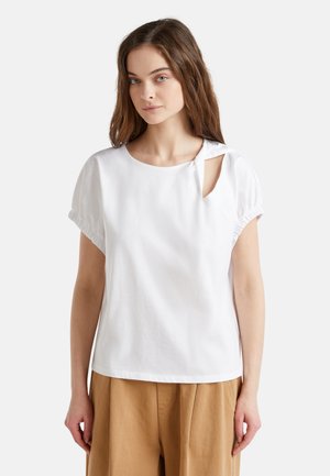 T-shirt en coton blanc avec un col rond, des manches courtes bouffantes et un détail découpé sur le côté. Il est associé à un pantalon large beige.