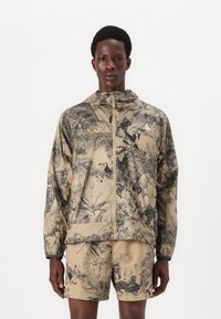 Beige Kapuzenjacke mit schwarzem botanischen Muster; verfügt über einen Reißverschluss und zwei Taschen, kombiniert mit passenden Shorts im selben Design.