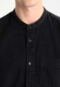 Chemise noire à col mandarin, avec une poche poitrine et des boutons noirs classiques sur un tissu lisse.