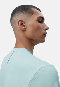 Svetlo modra teksturirana športna majica s krognim izrezom. Ima majhen navpični sivi poudarek na hrbtni strani blizu vratu. Gladek material.