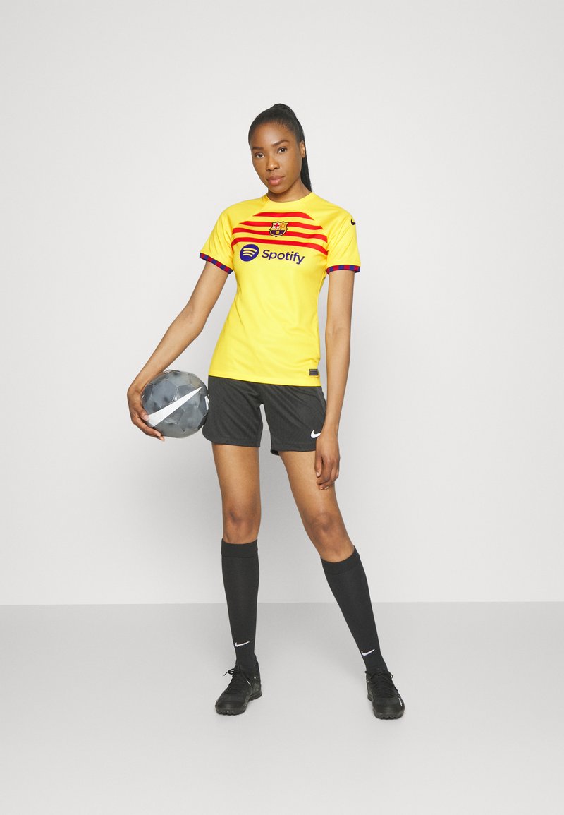 Camiseta de fútbol amarilla con rayas rojas y un logo, combinada con pantalones cortos negros y calcetas negras hasta la rodilla. Sosteniendo un balón de fútbol gris.