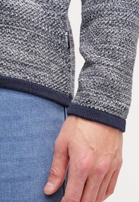 Šedý pletený svetr s texturovaným povrchem, s tmavě modrým žebrovaným lemem a manžetou, na boku se štítkem.