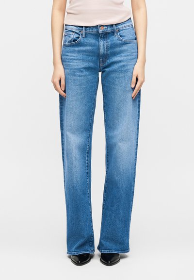 Γυναίκα με μπλε τζιν κανονικής γραμμής και μαύρα μυτερά παπούτσια, στέκεται με τα χέρια χαλαρά δίπλα στο σώμα μπροστά σε απλό φόντο.