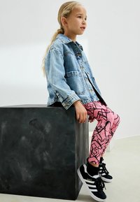 Jeansjacka, rosa leggings med svarta abstrakta mönster och svarta sneakers med vita sulor. Barn sitter på en grå, kubisk pedistal.
