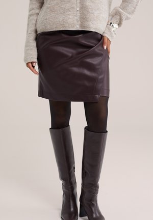 Vrouw die een donkerbruine leren minirok draagt met zwarte panty's, donkerbruine leren kniehoge laarzen en een lichtgrijze gebreide trui.