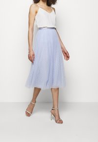 Kvinna i vit ärmlös topp, ljusblå midiklänning i tyll med prickmönster och silverhöga sandaler med ankelremmar.