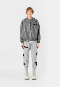 Cazadora bomber de cuero gris con cremallera frontal y bolsillos laterales, combinada con pantalones de chándal gris claro con patrones de aplique en blanco y negro. Botas blancas.