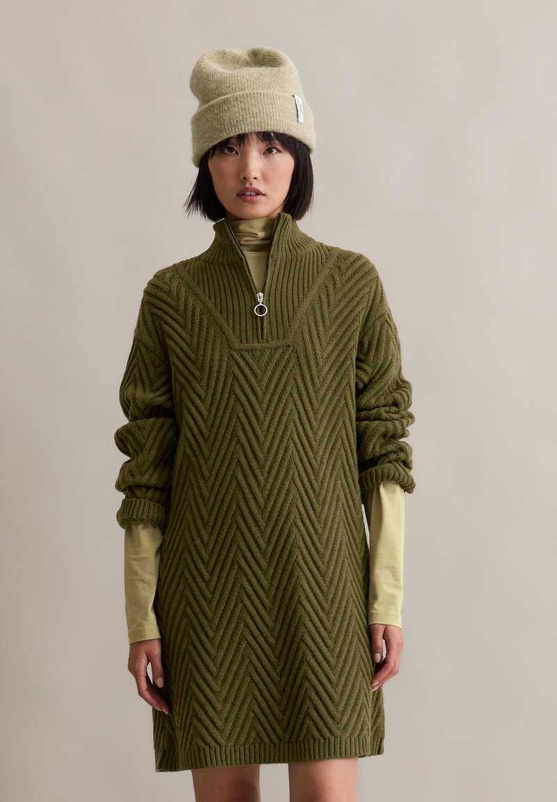 Robe en tricot vert olive avec un col montant et une texture en zigzag. À associer avec un bonnet vert clair. Manches longues et design décontracté.