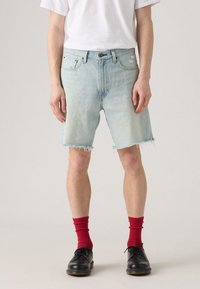 Shorts en denim bleu clair avec des bords effilochés, un design à cinq poches et une coupe décontractée, portés avec des chaussettes rouges et des chaussures noires.
