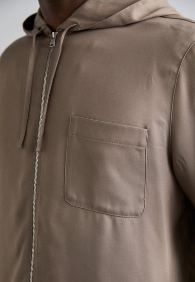 Φωτεινό καφέ μπουφάν με κουκούλα από απαλό ύφασμα, με φερμουάρ μπροστά και μια πλαϊνή τσέπη αριστερά. Μινιμαλιστικός σχεδιασμός χωρίς σχέδια.