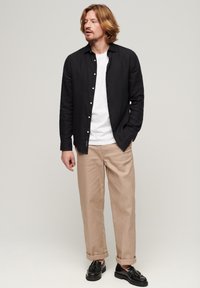 Camisa negra de lino de botones sobre una camiseta blanca, combinada con pantalones beige claros y zapatos negros. Diseño simple, ajuste casual.
