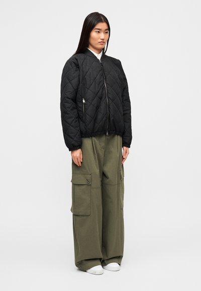 Μαύρο μπουφάν bomber με καπιτονέ μοτίβο, κλείσιμο με φερμουάρ και ριμπ μανσέτες, συνδυασμένο με φαρδιά χακί παντελόνια cargo με πολλαπλές τσέπες.