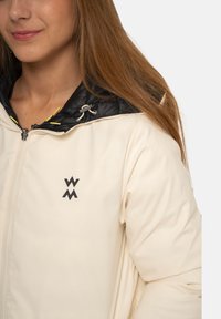 Veste de couleur crème avec une capuche isolée noire. Elle dispose d'une fermeture éclair à l'avant et d'un logo sur la poitrine. Matière lisse avec un léger éclat.