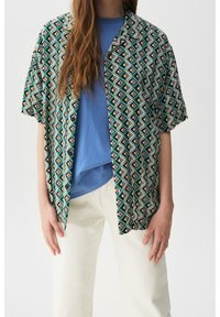 Chemise à manches courtes à motifs avec un design géométrique en vert, noir et beige, associée à un haut bleu et un pantalon couleur crème.