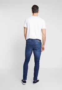 Jeans de denim ajustados en azul oscuro, con dos bolsillos traseros. Combinados con una camiseta blanca básica y zapatillas negras. Textura suave.