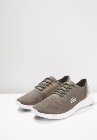 Chaussures de sport en maille marron à finition mate, avec des accents verts et des semelles en caoutchouc blanches. Les lacets sont verts assortis avec un marquage minimal.