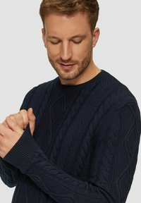 Mann mit kurzen braunen Haaren und Bart, der in einem dunkelblauen, grobstrickigen Sweater nach unten schaut, mit in der Brust gegeneinandergelegten Händen.