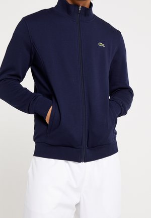 Mann trägt eine marineblaue Jacke mit Stehkragen, Seitentaschen und kleinem grünem Krokodillogo auf der Brust, kombiniert mit weißen Hosen.