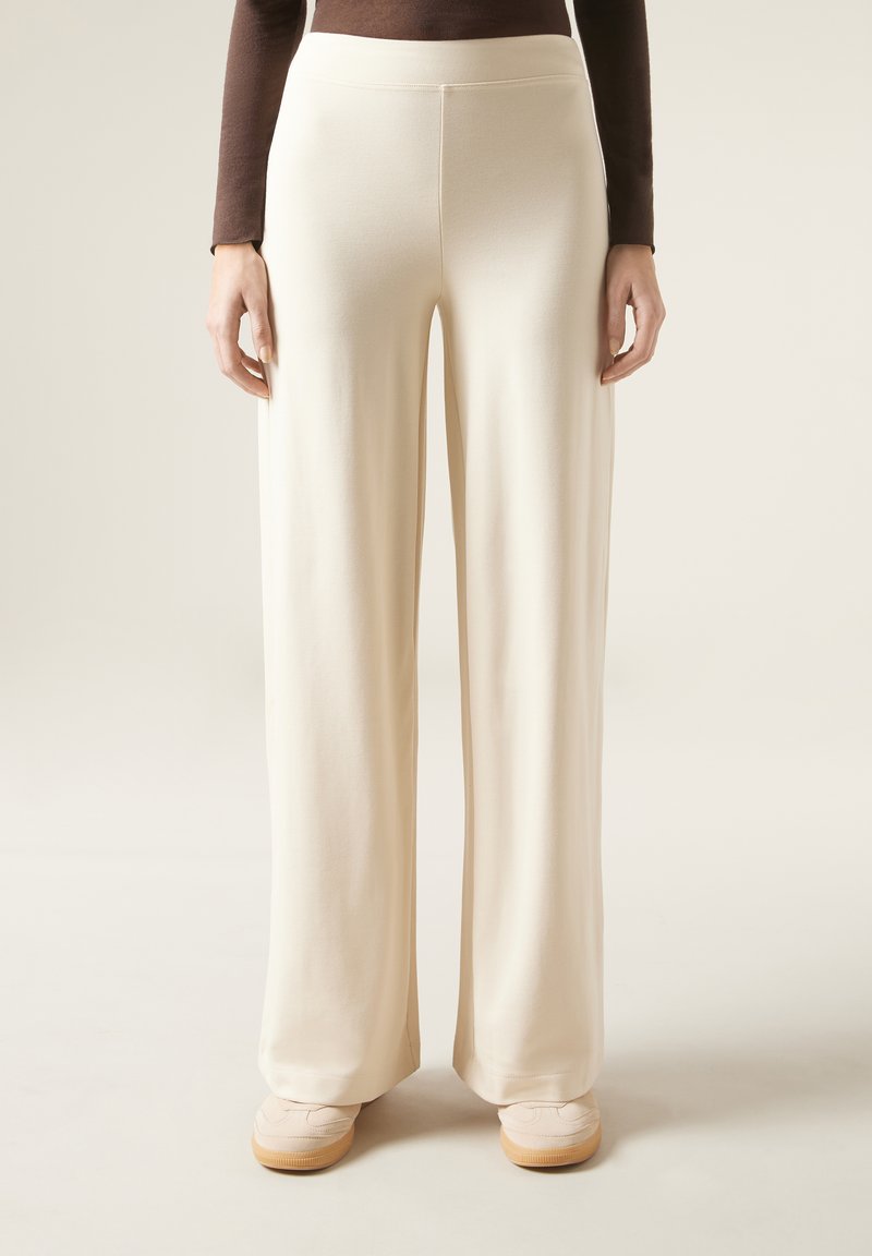 Pantalons à jambes larges en tissu beige clair et doux, présentant une texture lisse et un design taille haute, associés à des chaussures de couleur claire.