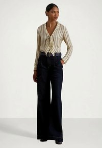 Γυναίκα ντυμένη με μια κρέμα και μαύρη κάθετα ριγέ μπλούζα με ζωνάκι, ψηλής μέσης ανοιχτόχρωμες φαρδιές τζιν παντελόνες και μαύρη ζώνη, στέκεται με τα χέρια στα τσέπες.