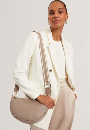 Nainen, jolla on valkoinen bleiseri ja beiget housut, kantaa olkapäällään taupen väristä puoliympyrän muotoista laukkua, jossa on merkitty olkahihna.