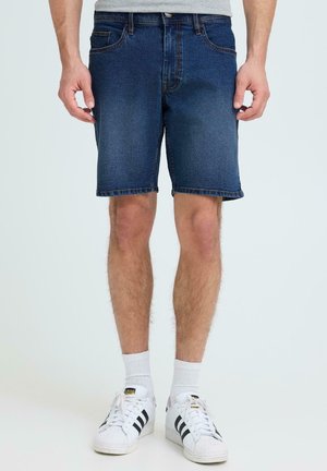 Homme portant un short en denim bleu, un t-shirt gris, des chaussettes blanches et des baskets Adidas blanches à rayures noires, debout devant un fond uni.