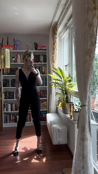 Macacão preto com decote em V profundo, cintura ajustada e pernas afuniladas. Usado com saltos pretos. Interior brilhante com estantes e plantas.