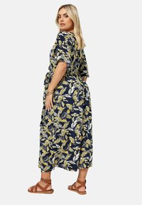 Blommig omlottklänning i marinblått med gula och vita bladmönster. Korta ärmar, midjehögt skärp och ankellång nederkant. Matchad med bruna sandaler.