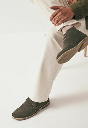 Pantofi din suede verde închis, cu un profil jos, având două găuri pentru stinghii, talpă din cauciuc plată și accente maro deschis aproape de talpă.