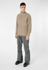 Beige langärmliges Zip-up Oberteil und graue Hose, mit strukturiertem Stoff und schlanker Passform. Schwarze Skistiefel runden das Outfit ab.