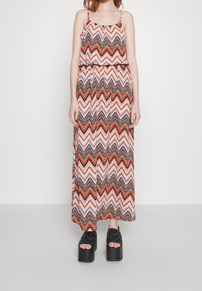 Vrouw draagt een mouwloze maxi-jurk met een zigzagpatroon in oranje, zwart en roze, gecombineerd met zwarte sleehakken.