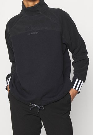 Osoba nosící černý fleecový pulovr Adidas s bílými pruhovanými manžetami a černé kalhoty, ruce v kapsách, na jednobarevném pozadí.