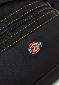 Melnas auduma mugursomas ar rāvējslēdzēju un kontrastējošu dzeltenu šuvju apdari; iekļauta taisnstūra "Dickies" logotipa uzlīme.
