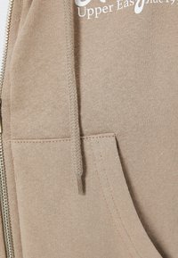 Felpa con zip beige, dotata di tasca frontale, coulisse e logo stampato. Tessuto di cotone liscio con dettagli di cucitura visibili.