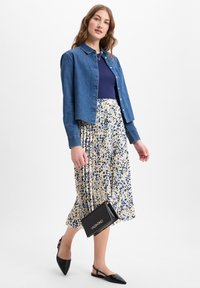 Jeansjacke in cropped-Form über einem marineblauen Hemd, kombiniert mit einem plissierten Blumenkleid in Creme, Blau und Schwarz. Schwarze spitze Ballerinas und eine kleine Handtasche.