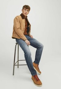 Chaqueta de pana color marrón claro con un cuello marrón, suéter color óxido, jeans de mezclilla azules y botas de senderismo marrones con cordones rojos, sentado en un taburete de metal.