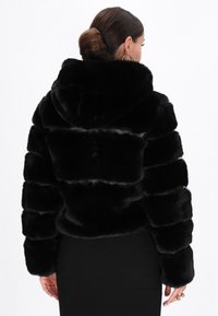 Sort faux fur jakke med hætte, med en quiltet design og lange ærmer. Teksturen er blød, med en glat og blank finish.
