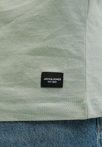 Világos zöld pamut póló, melynek bal alsó sarkában egy fekete címke található, rajta a "JACK & JONES EST. 1990" felirattal; farmernadrág látható alatta.