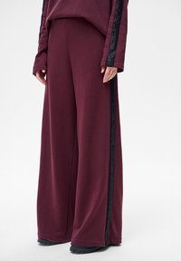 Wijde broek in bordeauxrode stof met een zachte textuur, voorzien van een zwarte, met patroon bedrukte zijstreep en een ontspannen pasvorm. Zwarte schoenen maken de look compleet.