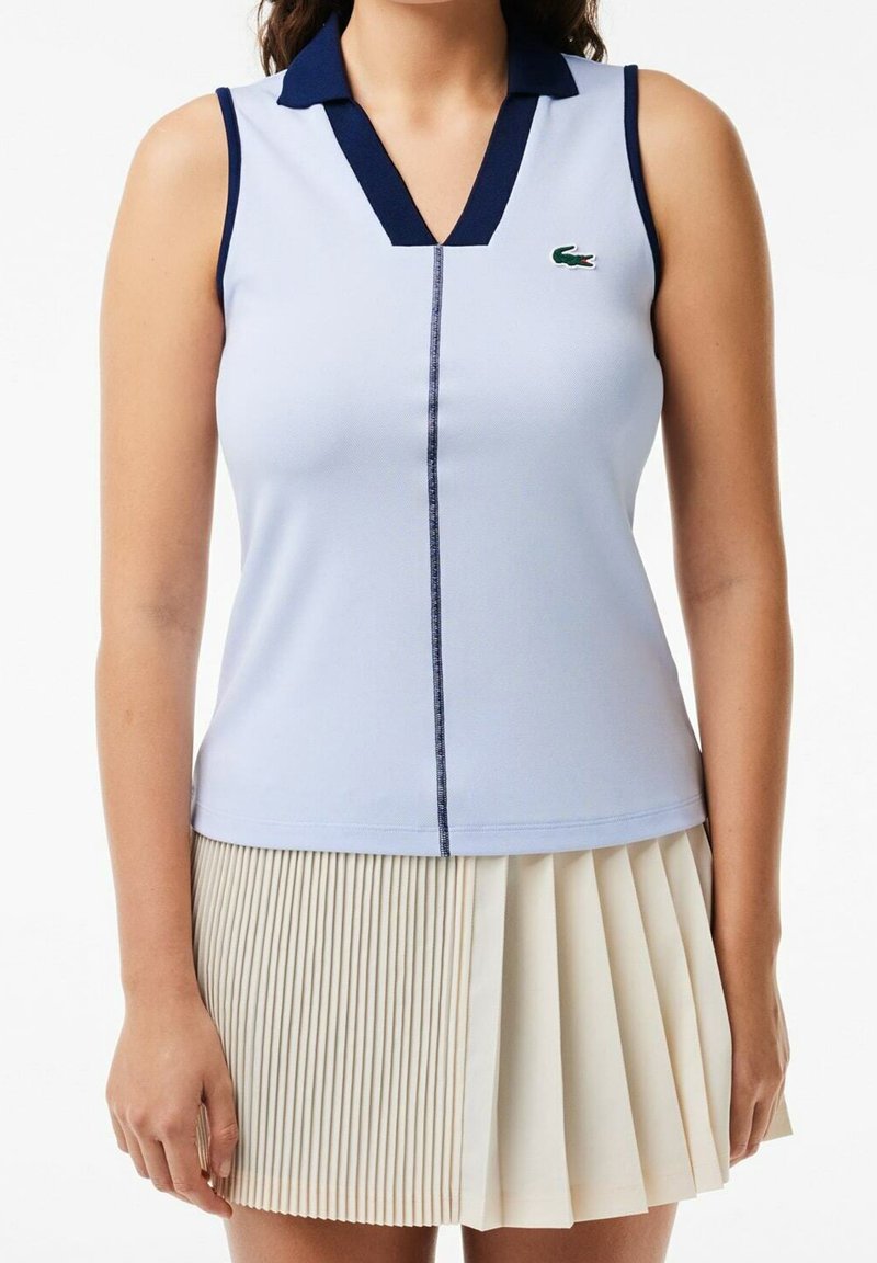 Haut bleu clair sans manches avec col bleu foncé et couture verticale, associé à une jupe plissée crème portée par une personne debout sur fond blanc.